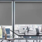 A meeting room with a large window covered by gray roller blinds, overlooking a city skyline.